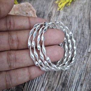Pode incluir: Um conjunto de pulseiras de metal prateado torcido. As pulseiras têm um design em espiral e são empilhadas. As joias são exibidas em uma superfície de madeira, com uma mão segurando as pulseiras.