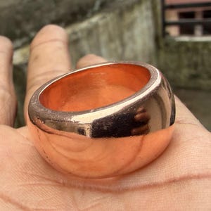 May include: A wide, polished copper ring with a smooth, rounded exterior surface and a metallic, reddish-brown finish. The band features a thick, solid construction with a reflective sheen that catches the light across its curved, circular form.