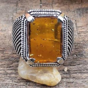 May include: A silver ring featuring a rectangular amber-coloured stone. The ring has a detailed, textured band and is set on a small, light-coloured stone against a wooden surface. The amber stone has a warm, golden hue.