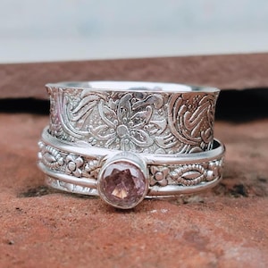 May include: A silver-colored ring with a floral design and a pink gemstone. The ring has a spinning band with an embossed floral pattern. The gemstone is round and set in a decorative bezel. The ring is on a reddish-brown surface.