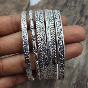 Puede incluir: Un conjunto de seis brazaletes de metal plateado con varios patrones en relieve, incluyendo motivos florales, formas ovaladas repetitivas y texturas trenzadas. Cada pulsera tiene un diseño distinto con detalles oxidados en las áreas.