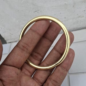 以下が含まれることがあります： 金色の円形バングルブレスレット。滑らかで磨かれた表面が光を反射し、明るく光沢のある外観を作り出しています。バングルはシンプルでエレガントなデザインで、普段使いに適しています。ブレスレットの直径は約7.6cmです。