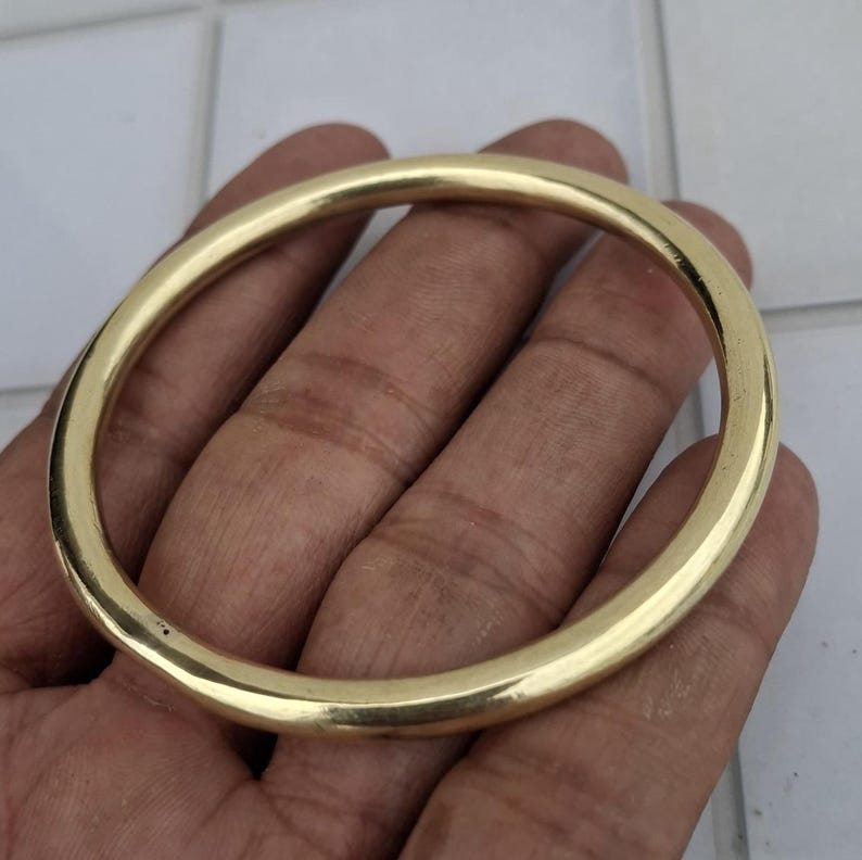 以下が含まれることがあります： 金色の円形バングルブレスレット。ブレスレットは、滑らかで光沢のある表面を持つ、金属製のソリッドバンドです。バングルは手で持たれており、その丸い形と金属的な輝きを際立たせています。ブレスレットはシンプルでエレガントなジュエリーです。