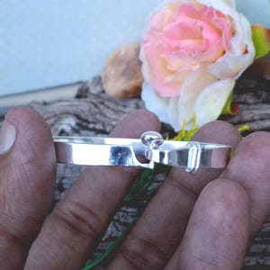 Könnte beinhalten: Ein silbernes Armband mit einem einzigartigen Verschluss. Das Armband hat ein rechteckiges Band mit einem kleinen, runden, erhabenen Element und einem hakenförmigen Verschluss. Das Design ist schlicht und elegant, geeignet für den täglichen Gebrauch.