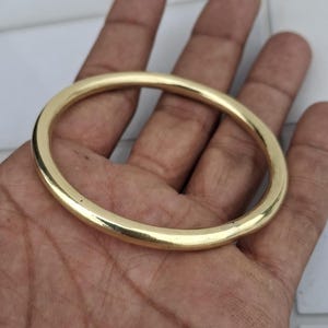 以下が含まれることがあります： 金色の楕円形のブレスレット。ブレスレットは滑らかで光沢があり、丸みを帯びた形状をしています。このジュエリーは人の手で持たれており、その研磨された表面とシンプルなデザインが際立っています。このブレスレットは、クラシックなジュエリーです。