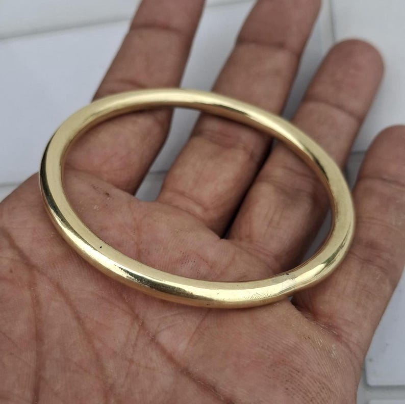 以下が含まれることがあります： 金色の楕円形のブレスレット。ブレスレットは滑らかで光沢があり、丸みを帯びた形状をしています。このジュエリーは人の手で持たれており、その研磨された表面とシンプルなデザインが際立っています。このブレスレットは、クラシックなジュエリーです。