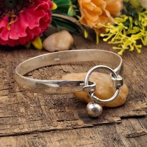 May include: A silver bangle bracelet featuring a circular clasp and a small, dangling ball. The bracelet is displayed on a small, light-coloured stone, with flowers in the background. The bracelet is made of a shiny metal.