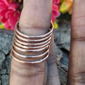 May include: A collection of copper rings worn on a finger. The rings are a warm, metallic colour and have a simple, minimalist design. The background features blurred pink and yellow flowers.