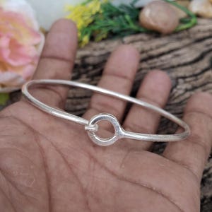 Könnte beinhalten: Ein silbernes Armreif-Armband mit einem kreisförmigen Verschluss. Das Armband ist dünn und hat ein schlichtes, elegantes Design. Der Verschluss ist ein kleiner, offener Kreis. Das Armband wird auf einer Hand gezeigt.