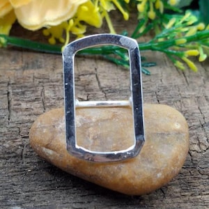May include: A silver-colored ring with a rectangular design. The ring is set on a smooth, light brown stone, with a wooden background and some greenery in the background.