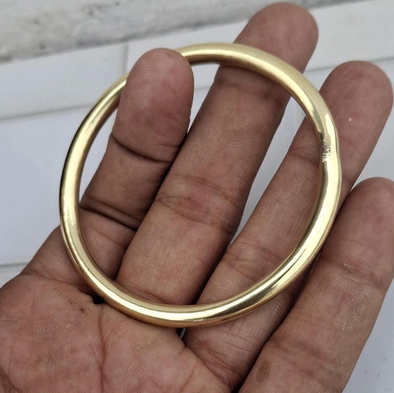 以下が含まれることがあります： 滑らかで丸みを帯びたデザインの、金色のバングルブレスレット。 ブレスレットは人の手で持たれており、その磨かれた表面と円形を示しています。 このジュエリーは、シンプルでありながらエレガントなアクセサリーです。