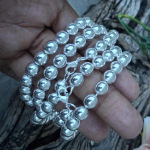 May include: A set of three silver beaded bracelets with a lobster clasp closure. The bracelets are made of shiny, round silver beads. The bracelets are displayed on a wooden surface.