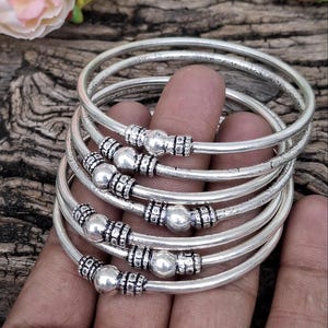 May include: A stack of silver bangles with decorative beads and etched detailing. The bracelets are round and have a polished finish. The bangles are displayed on a wooden surface.