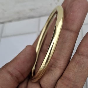 以下が含まれることがあります： 滑らかで丸みを帯びたデザインの、金色のバングルブレスレット。 ブレスレットは光を反射する研磨仕上げです。 このジュエリーはシンプルでエレガントなアクセサリーです。