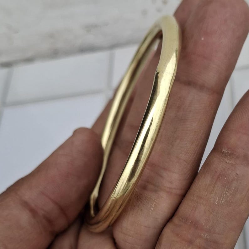 以下が含まれることがあります： 滑らかで丸みを帯びたデザインの、金色のバングルブレスレット。 ブレスレットは光を反射する研磨仕上げです。 このジュエリーはシンプルでエレガントなアクセサリーです。
