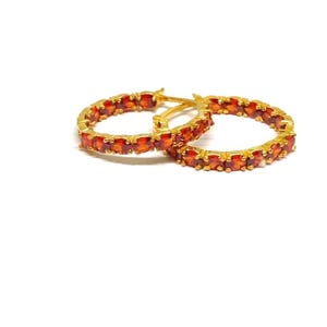 May include: A pair of gold-colored hoop earrings. Each earring is adorned with a row of small, oval-shaped, orange-red gemstones. The earrings are set against a white background.