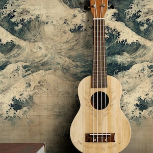 May include: A light-colored ukulele stands against a backdrop of a wave-patterned wallpaper. Two books rest on a dark wooden table in the foreground. The ukulele has four strings and a round sound hole.