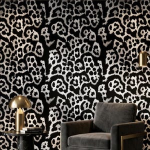 Puede incluir: Un papel pintado con estampado de leopardo en blanco y negro cubre la pared detrás de un sillón gris y una pequeña mesa auxiliar con una lámpara dorada. Una segunda lámpara dorada está a la derecha. La habitación está iluminada por luces empotradas en el techo.