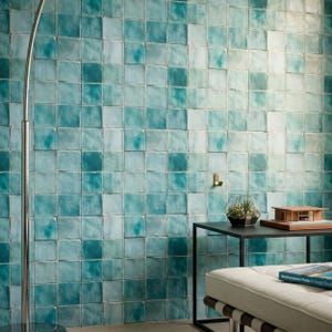 May include: A wall covered in square, glossy tiles featuring a gradient of turquoise, teal, and light blue hues with subtle variations in texture. A tall, thin, silver metal floor lamp stands to the left, and a black table holds a small plant.