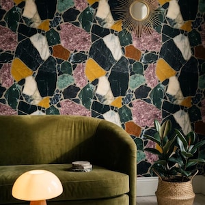 May include: A living room scene featuring a green velvet sofa, a round terrazzo coffee table, and a mushroom-shaped lamp. The wall is adorned with a colorful, abstract mosaic wallpaper. A gold sunburst mirror hangs on the wall, and a potted plant sits in a woven basket.