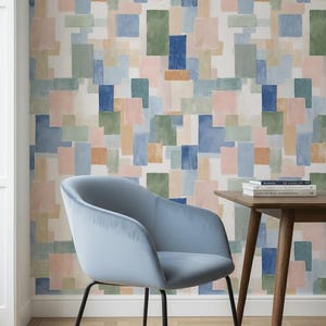 Op de afbeelding: Een lichtblauwe fluwelen fauteuil met zwarte metalen poten staat voor een muur bedekt met een aquarelpatroon van pastelkleurige rechthoeken. Een kleine houten tafel met boeken staat ernaast.