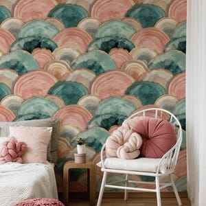 May include: A bedroom scene featuring a watercolor-style wallpaper with a repeating pattern of layered, semi-circular shapes in shades of pink, teal, and beige. A white chair with pink pillows, a bed with pink pillows, and a knitted ottoman are in the foreground.