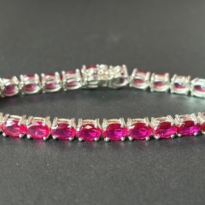 Könnte beinhalten: Ein silberfarbenes Tennisarmband mit einer durchgehenden Reihe oval geschliffener, leuchtend pinker Steine, die in einzelnen Metallfassungen sitzen. Das Armband verfügt über einen sicheren Kastenschloss-Verschluss in der Mitte.