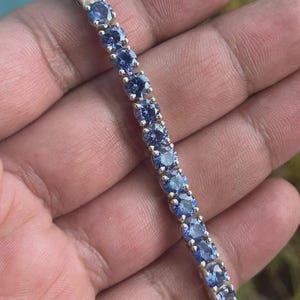 以下が含まれることがあります： 丸い青色の宝石が連なったシルバーブレスレット。宝石は互いに密接にセットされ、連続した色のラインを形成しています。ブレスレットはニュートラルな背景に表示され、石の輝きを際立たせています。