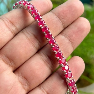Puede incluir: Una pulsera de plata con una fila de gemas rojas redondas y vibrantes. Las gemas están engarzadas juntas, creando una banda continua de color. La pulsera se sostiene en una mano, mostrando su diseño y brillo.