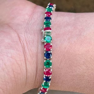 May include: A tennis bracelet with alternating red, green, and blue gemstones. The bracelet is made of silver and has a secure clasp. The gemstones are round and set closely together, creating a continuous line of color.