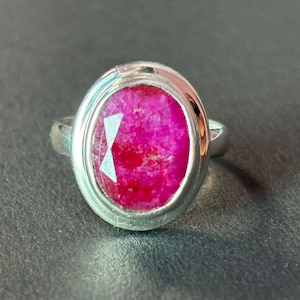 Könnte beinhalten: Ein Silberring mit einem großen, ovalen, leuchtend pinkfarbenen Edelstein, der in einer glatten, polierten Metallfassung sitzt. Der Stein hat eine facettierte Oberfläche, die das Licht reflektiert, und die Ringschiene ist schlicht und poliert.