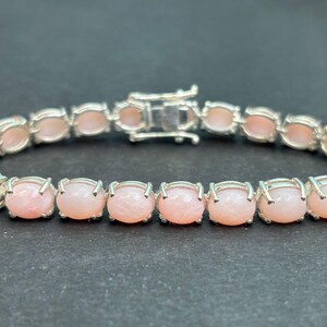 May include: A silver bracelet featuring a series of light pink, oval-shaped stones. Each stone is set in a silver bezel, and the bracelet is secured with a silver clasp. The stones are evenly spaced and create a continuous line.