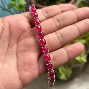 Op de afbeelding: Een zilveren armband met ovale, levendige rode edelstenen. De edelstenen zijn in een rij geplaatst, waardoor een doorlopend patroon ontstaat. De armband wordt getoond tegen een neutrale achtergrond, wat de kleur en het ontwerp benadrukt.