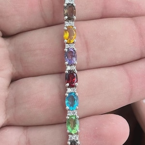 May include: A silver bracelet featuring a series of oval-cut gemstones in various colors, including blue, green, yellow, and purple. Small, clear stones are interspersed between the larger gems. The bracelet is held against a blurred background.