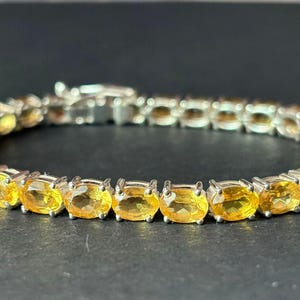 May include: A silver bracelet with a row of oval, yellow gemstones. The stones are set in individual prongs, and the bracelet fastens with a clasp. The bracelet is shown against a dark backdrop.