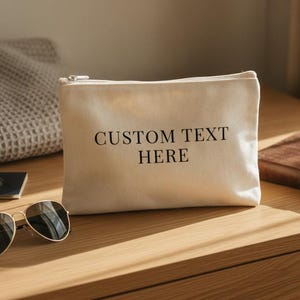 Op de afbeelding: Een rechthoekig, gebroken wit canvas etui met ritssluiting en de in het zwart gedrukte tekst CUSTOM TEXT HERE gecentreerd op de voorzijde. Het etui is voorzien van een zilverkleurige metalen ritssluiting aan de bovenzijde.