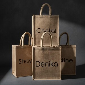 May include: A collection of four rectangular tan jute tote bags with two top handles each. The bags are arranged in a stack, with each bag featuring a black printed name on the front. The visible names are Crystal, Shay, Denika, and tine.