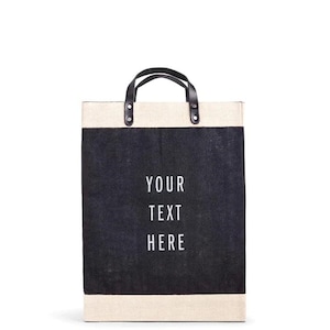 Op de afbeelding: Een rechthoekige draagtas met een zwart middenpaneel en crèmekleurige boven- en onderdelen. De tas heeft zwarte leren handvatten en de tekst "YOUR TEXT HERE" in het wit.