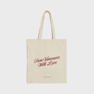 Op de afbeelding: Een beige canvas draagtas met twee schouderbanden. De voorzijde is voorzien van de donkerrode cursieve tekst From Vancouver With Love en de kleinere donkerrode tekst PIPPA + OLIVER onderaan in het midden van de tas.