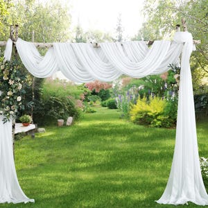 May include: A white fabric wedding arch with a wooden frame, draped with flowing white fabric. The arch is decorated with floral arrangements and stands on a green lawn, creating a romantic outdoor setting.