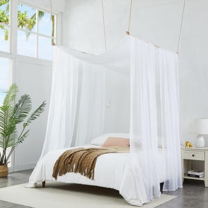 May include: A white four-poster bed frame with sheer white fabric curtains draped from the top rails. The bed is dressed in white bedding with a textured brown throw blanket folded at the foot. The frame is suspended by thin ropes from the ceiling.