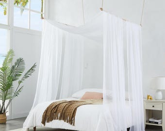May include: A white canopy bed with sheer curtains suspended from a wooden frame. The bed is made with white linens and a brown throw blanket. A white nightstand with a lamp and a small gold figurine sits beside the bed.