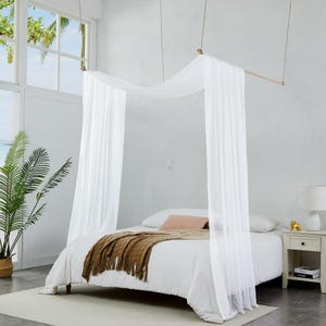 Op de afbeelding: Een wit hemelbed met transparante, vloeiende stof die aan een houten staaf is opgehangen. Het bed is opgemaakt met wit beddengoed en een bruine deken. Een wit nachtkastje met een lamp en decoratie staat naast het bed.
