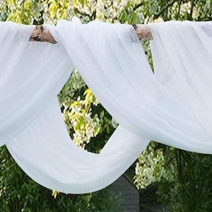 以下が含まれることがあります： 天然の木の枝に白いシアー生地をドレープし、流れるようなエレガントなデザインを作り出しています。生地は柔らかい折り目で配置され、背景にはぼやけた緑の葉があります。