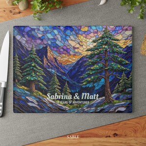May include: A rectangular cutting board with a stained glass design featuring a mountain landscape with pine trees. The text "Sabrina & Matt 10 YEARS OF ADVENTURES" is displayed on the bottom. The board is on a gray surface.