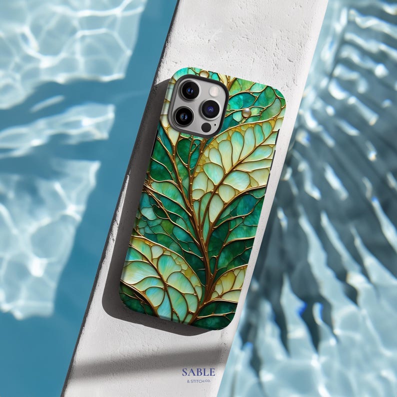 May include: A smartphone case with a vibrant leaf design. The case features a pattern of green and gold leaves with intricate details. The case is resting on a white surface with a blue water background. The text "SABLE & STITCH CO." is visible.