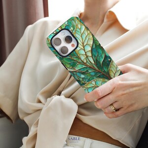 May include: A smartphone case with a green and gold leaf design. The case features a detailed, artistic rendering of a leaf with intricate veining and a metallic gold outline. The case is held in a person's hand.