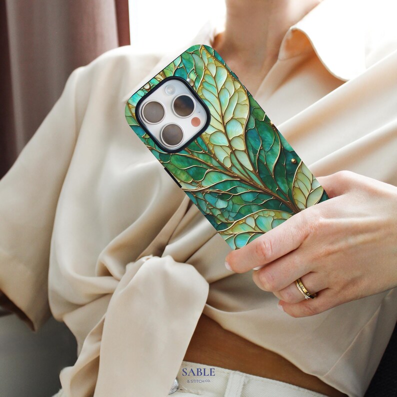 May include: A smartphone case with a green and gold leaf design. The case features a detailed, artistic rendering of a leaf with intricate veining and a metallic gold outline. The case is held in a person's hand.