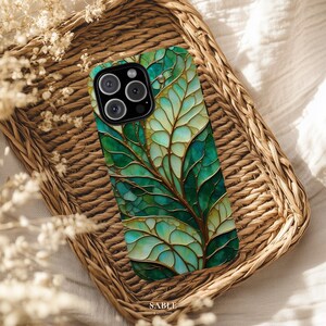 May include: A smartphone case with a green and gold leaf design, resting on a woven tray. The case features a detailed illustration of leaves in shades of green and gold, with a dark camera module. The tray is made of natural woven material. The brand name "SABLE" is visible.
