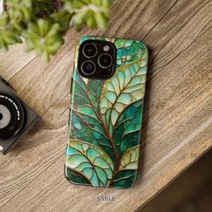 May include: A smartphone case with a green and gold leaf design. The case features a detailed, stained-glass-like pattern with shades of green and gold. The word "SABLE" is printed at the bottom of the case.